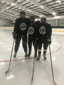 3 Hockey Players in full hockey gear stand on the ice with their hockey sticks.Their jersey’s read “NY Metro Blind Hockey”