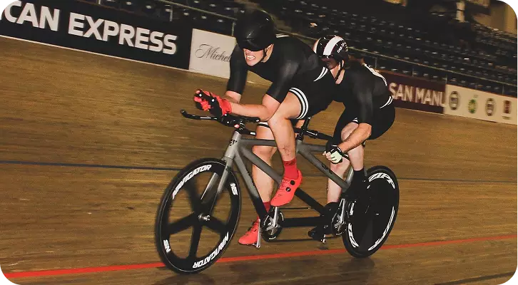 Two male cyclists racing at high speed on a grey tandem bike around a wooden velodrome track. The sighted pilot in front and the visually impaired athlete in the back are wearing matching black skinsuits and aerodynamic helmets, leaning intensely into a turn.