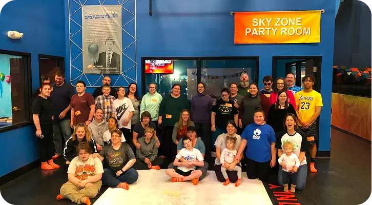 A large, smiling group of visually impaired youth, their families, and volunteers posing for a group photo inside the 'Sky Zone Party Room'. Participants of all ages are gathered together against a blue wall, wearing orange trampoline socks, representing a vibrant community event.