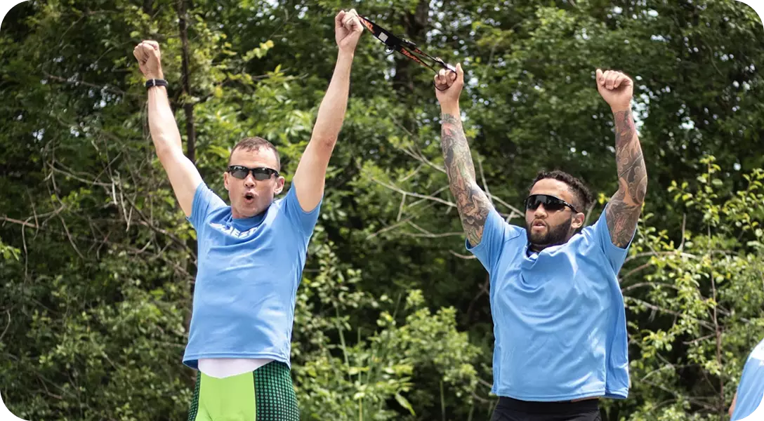 Two athletes, likely a visually impaired participant and his guide, celebrating a moment of triumph outdoors. Both men are wearing matching light blue t-shirts and sunglasses, raising their arms high in the air with expressions of pure joy and accomplishment against a backdrop of green trees.