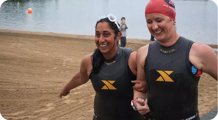 Two female open-water swimmers, a visually impaired athlete and her sighted guide, walking arm-in-arm across a sandy beach after exiting the water. Both are wearing black wetsuits and smiling broadly, demonstrating the strong bond of trust and partnership found in adaptive sports.