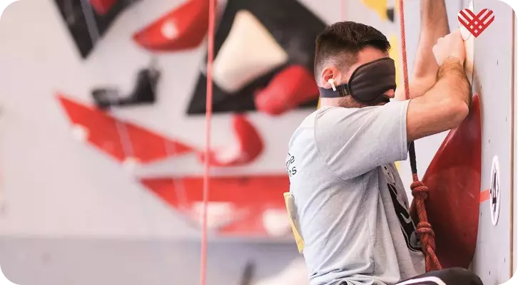 A male climber scaling an indoor wall while wearing a black eye mask to simulate total blindness or ensure fairness in competition. He is wearing a white earbud to listen to verbal directions from a 'caller' on the ground, highlighting the importance of auditory communication and trust in blind climbing.