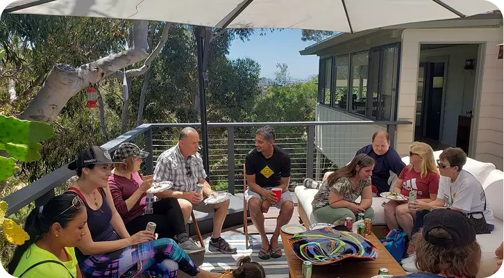 A casual social gathering of adults, including community members and volunteers, sitting in a circle on a sunny outdoor deck under a large white umbrella. They are enjoying a meal and engaging in conversation, highlighting the importance of peer support and social connection within the foundation.