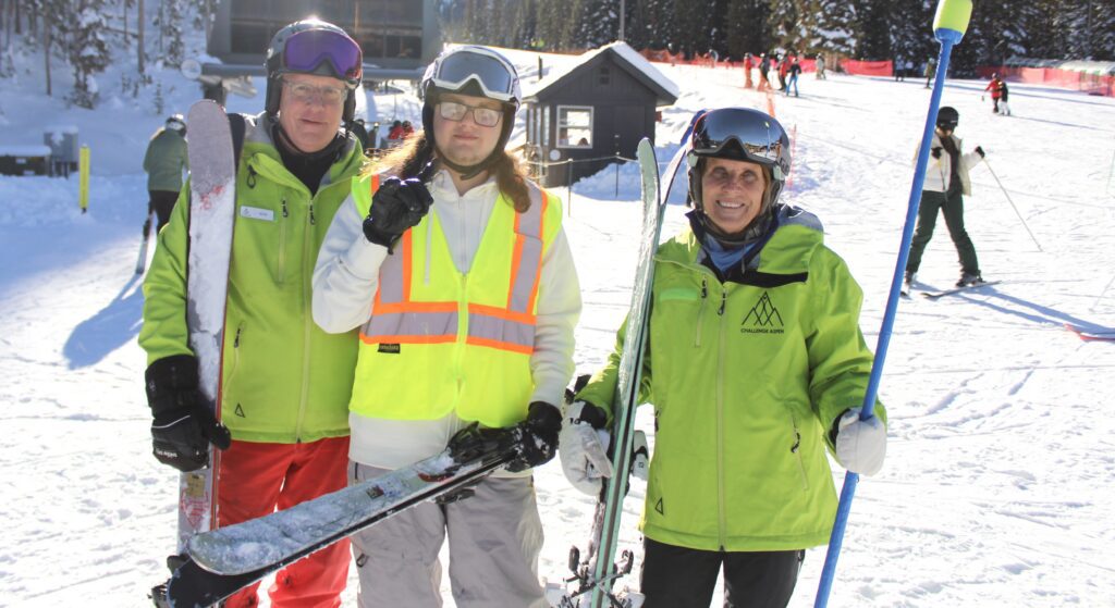 A male student is standing between a male guide and female guide on the ski slopes.