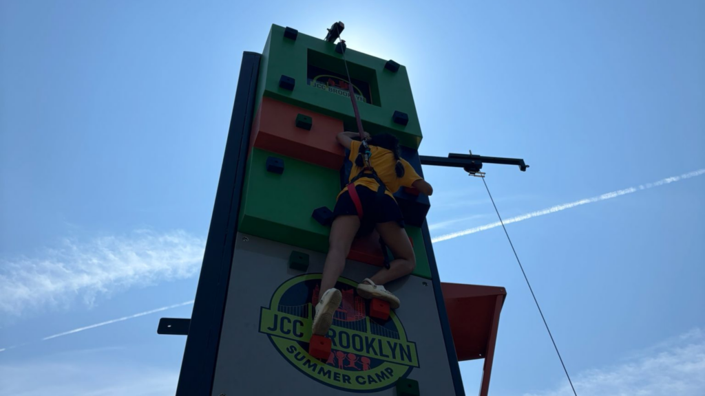 In this photo, a young girl is climbing an outdoor rock walk with an auto-belay.