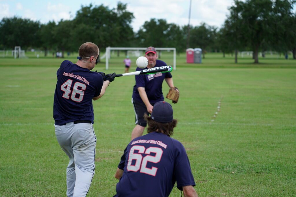 A legally blind man is holding a baseball bat about to hit a baseball.
